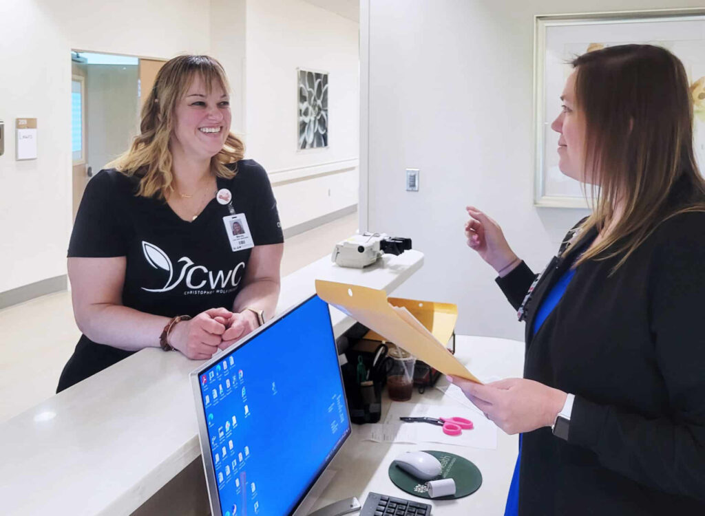 Two individuals interacting at a hospital reception desk. One person is standing behind the desk, smiling and wearing a black top with a badge, while the other, holding a manila folder, is engaging in conversation. Visible on the desk are a computer, a telephone, and various office supplies.