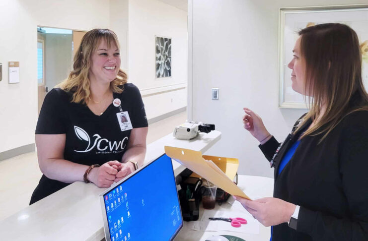 Two individuals interacting at a hospital reception desk. One person is standing behind the desk, smiling and wearing a black top with a badge, while the other, holding a manila folder, is engaging in conversation. Visible on the desk are a computer, a telephone, and various office supplies.