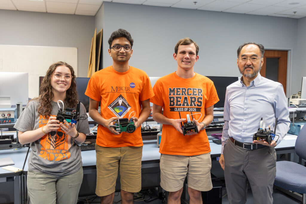 Three Mercer University students and a professor in a laboratory holding small robots. The laboratory setting includes various pieces of equipment and computer monitors in the background.