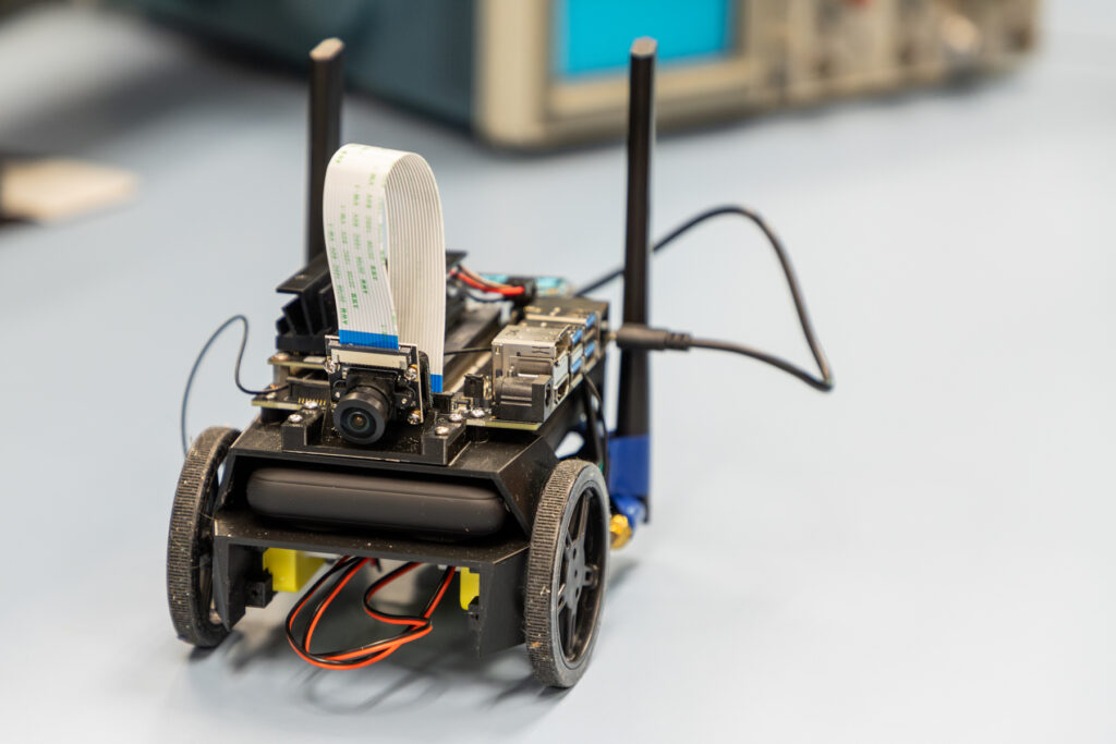 A small, wheeled robot is shown on a table. The robot features a camera mounted on top with a flat ribbon cable, two antennas, various electronic components, and visible wiring.