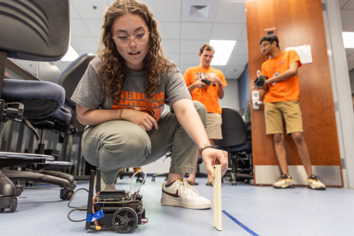 A Mercer University student crouches on the ground holding a small panel of wood in front of a small, wheeled robot.