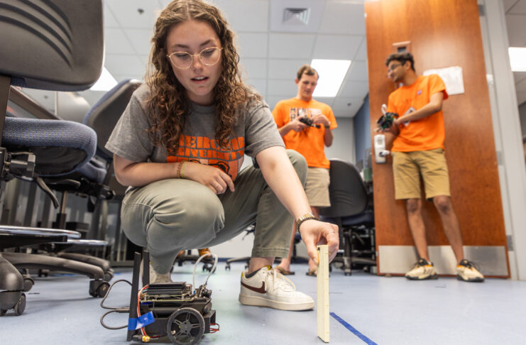 A Mercer University student crouches on the ground holding a small panel of wood in front of a small, wheeled robot.