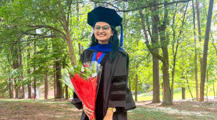 Graduate Spotlight: Priyal Bagwe A young woman stands outdoors in academic regalia, holding a bouquet of flowers. She wears a black graduation gown with velvet panels, a blue hood, and a black tam (a soft, round cap with a tassel). The setting is lush and green, with tall trees and sunlight filtering through the leaves. She smiles brightly at the camera.
