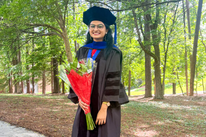 A young woman stands outdoors in academic regalia, holding a bouquet of flowers. She wears a black graduation gown with velvet panels, a blue hood, and a black tam (a soft, round cap with a tassel). The setting is lush and green, with tall trees and sunlight filtering through the leaves. She smiles brightly at the camera.