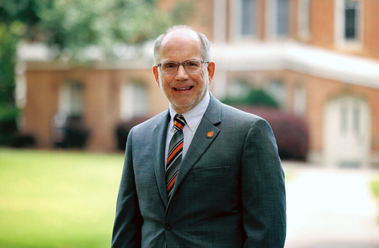 A man stands outside in front of a blurred brick building. He is wearing a gray suit, a striped tie in black, orange, and white, and glasses. He has a friendly smile and is slightly balding with a neatly trimmed gray beard. A circular orange and black pin is attached to his suit lapel.
