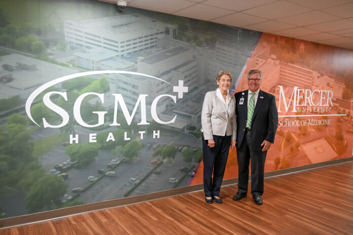 Mercer School of Medicine Dean Dr. Jean Sumner and SGMC Health CEO Ronnie Dean stand in front of a wall that is painted with a mural that has the words SGMC Health on the left and Mercer University School of Medicine on the right.