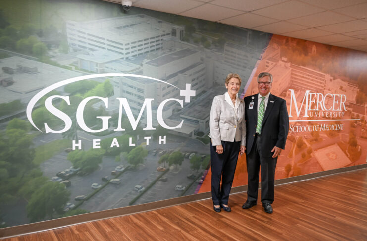 Mercer School of Medicine Dean Dr. Jean Sumner and SGMC Health CEO Ronnie Dean stand in front of a wall that is painted with a mural that has the words SGMC Health on the left and Mercer University School of Medicine on the right.