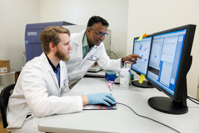 Dr. Raghavan Chinnadurai and biomedical sciences student working in a lab. The student controls a computer mouse, and Dr. Chinnadurai points at the computer monitor.