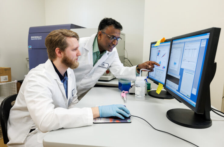 Dr. Raghavan Chinnadurai and biomedical sciences student working in a lab. The student controls a computer mouse, and Dr. Chinnadurai points at the computer monitor.