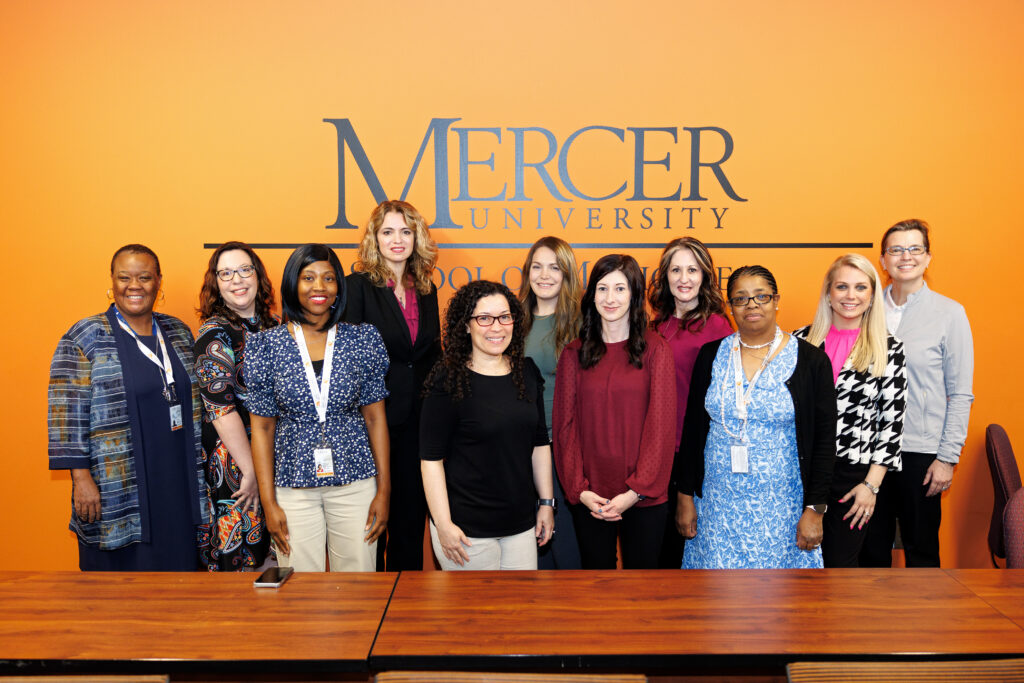 Group of 11 individuals standing in front of a wall with the “Mercer University” logo, smiling at the camera. They are in a professional setting, dressed in business attire. The background has an orange hue and the logo is in a dark blue font.