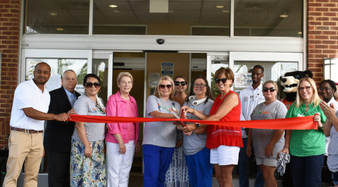 KidsABC celebrates Clinch Memorial Hospital’s Emergency Department as part of the Pediatric Emergency Care Project A group of people stand in front of a building entrance, holding a red ribbon for a ceremonial ribbon cutting. Some are wearing casual attire, while others are dressed more formally. There is a mascot in the background, and the mood is celebratory.