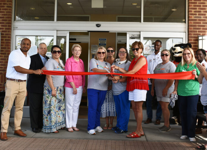 A group of people stand in front of a building entrance, holding a red ribbon for a ceremonial ribbon cutting. Some are wearing casual attire, while others are dressed more formally. There is a mascot in the background, and the mood is celebratory.