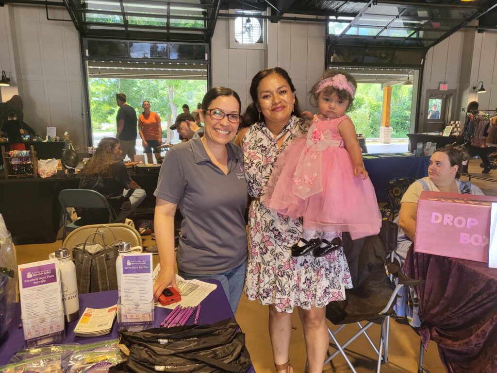 A woman wearing a South Georgia Healthy Start shirt stands next to a woman in a floral dress holding a baby girl in a pink tulle dress. They smile and pose together at a community health event, surrounded by informational booths and attendees.