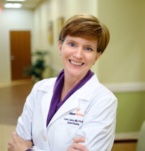 Dr. Lynn Denny wears a white coat and stands with her arms crossed in an office hallway.