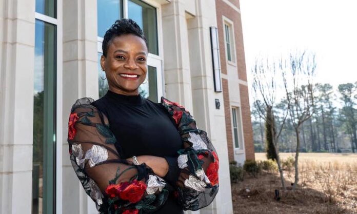 Dr. Pamela Moye stands in front of the College of Pharmacy. She is wearing a black top, her arms are crossed, and she is smiling.