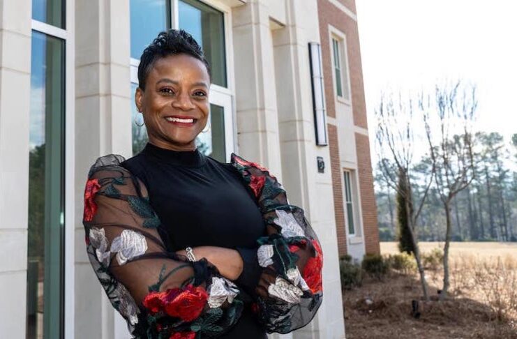 Dr. Pamela Moye stands in front of the College of Pharmacy. She is wearing a black top, her arms are crossed, and she is smiling.