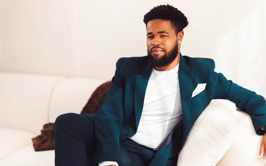 A man in a dark blue suit and white shirt and pocket square sits on a white couch, with his arm draped over one side.
