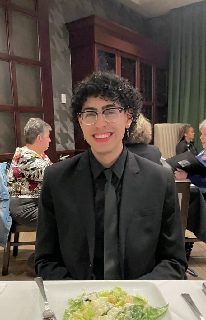 A man with curly hair and glasses wearing a black shirt, smiling at a dining table with a salad in front of them.