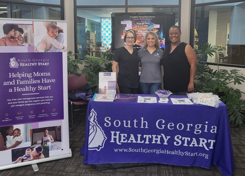 Four individuals stand behind a promotional booth for South Georgia Healthy Start inside a bright, airy atrium, surrounded by informational banners and leaflets related to maternal and family health.