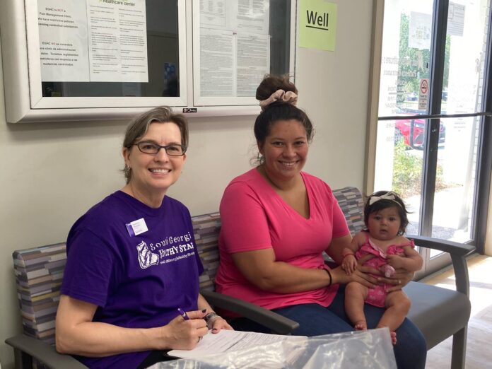 A woman in a South Georgia Healthy Start purple shirt sits on a bench with a woman in a bright pink shirt holding a baby girl in a pink onesie in a waiting room.