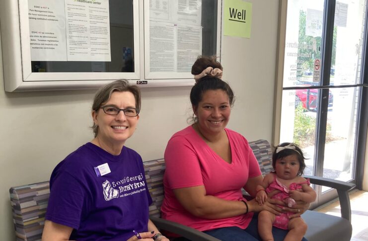 A woman in a South Georgia Healthy Start purple shirt sits on a bench with a woman in a bright pink shirt holding a baby girl in a pink onesie in a waiting room.