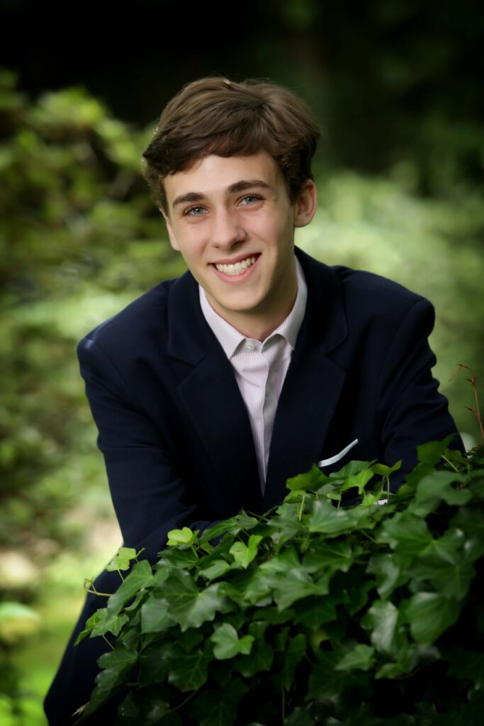 A young man in a dark blue suit and light pink shirt, smiling and leaning forward slightly, set against a blurred background of lush green foliage.