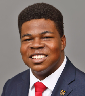 Headshot of Alex Marshall wearing a navy blue suit jacket, white collared shirt and red tie.