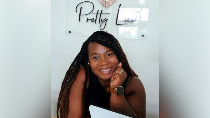 A women smiling at the camera with their chin resting on their hand, sitting at a desk with a laptop. Behind them, a wall sign reads "Pretty Lux."