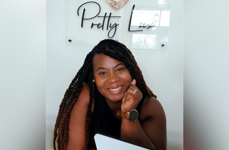 A women smiling at the camera with their chin resting on their hand, sitting at a desk with a laptop. Behind them, a wall sign reads 