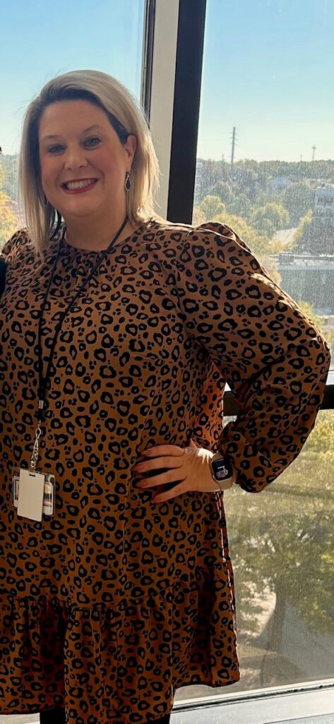 A woman in a leopard print dress smiling and posing confidently indoors by a window overlooking a tree-lined cityscape.