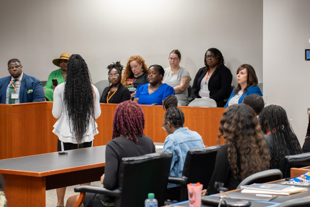 A diverse group of people sit in a jury box in a courtroom setting listening to a speaker who is standing.