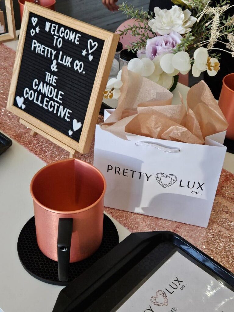 A promotional display for Pretty Lux Co., featuring a candle, a bag with a logo, and a welcome sign on a decorated table.