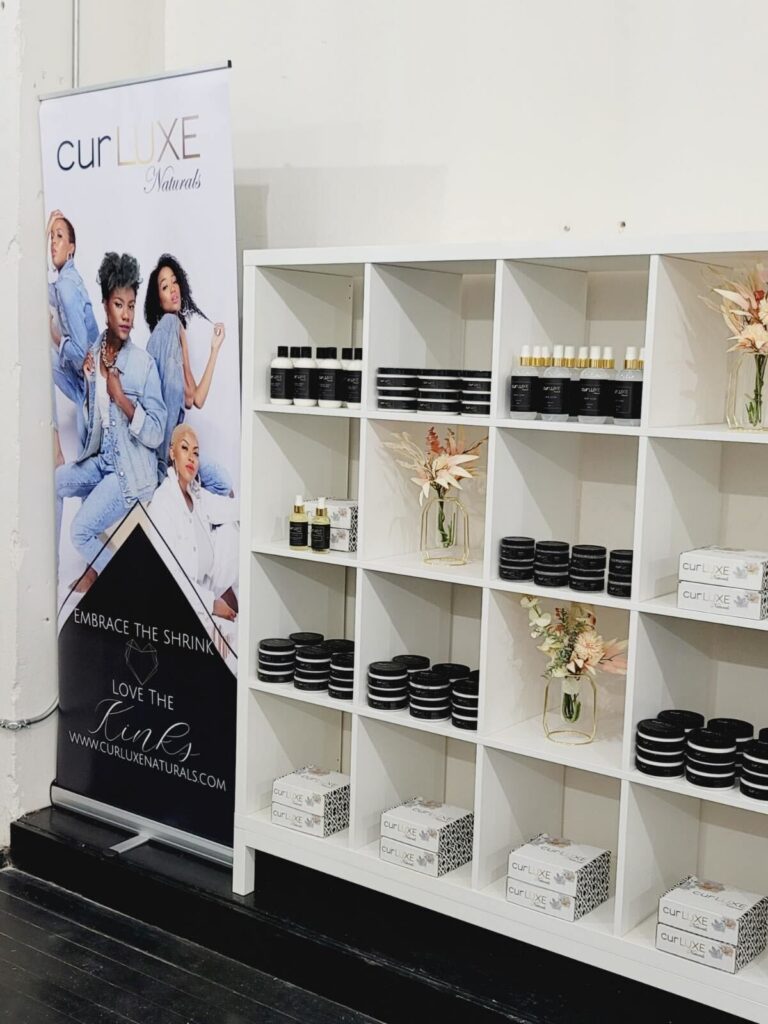 A retail display for CurLUXE Naturals products featuring neatly arranged beauty products on white shelves and a promotional banner with three models posing.