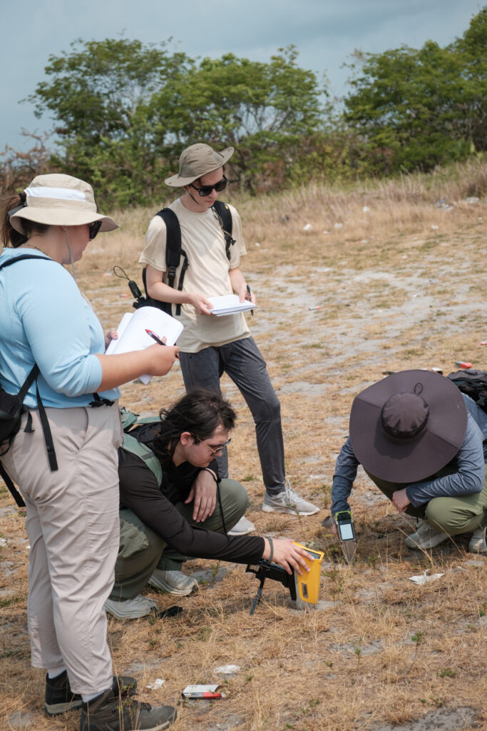 Four Mercer University students in a field, using equipment and taking notes. Two students are kneeling with their equipment pointed at the ground, while others observe and write in notebooks. They are dressed in casual field gear appropriate for outdoor research.