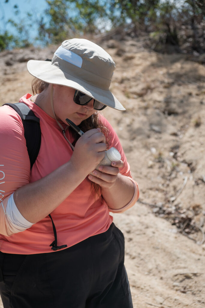 A Mercer University student writing on a small bottle filled with soil, wearing a sun hat and a backpack, outdoors on a sunny day.