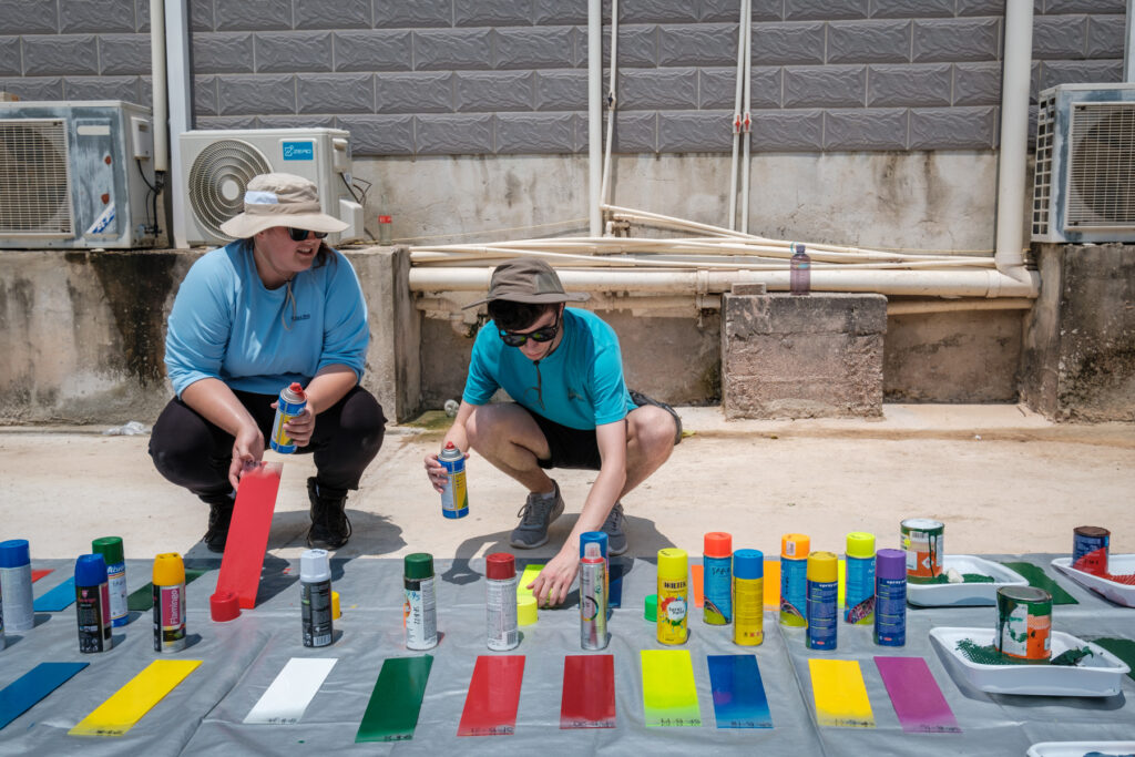 Two individuals, wearing caps and casual attire, hold cans of spray paint while more cans and painted color swatches are laid out on a tarp on the ground in front of them.