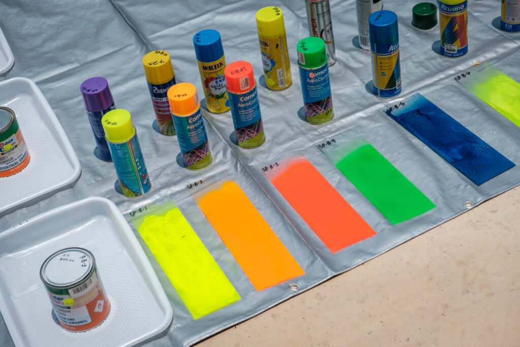 Multiple spray paint cans of colorful paint alongside corresponding painted color swatches on a plastic-covered surface. The colors include yellow, orange, red, green and blue. The cans are varied in brand and type, with labels visible.