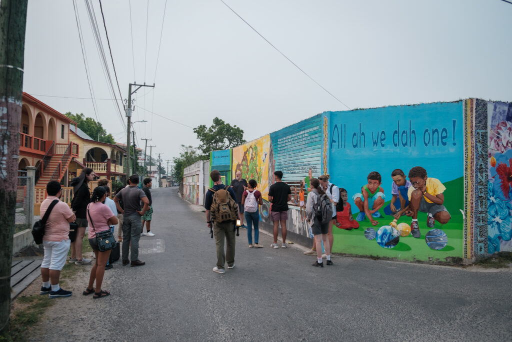 A group of people stand in a street alongside a colorful mural on a building wall depicting several children playing. The mural includes the text "All ah we dah one!" which suggests a theme of unity or togetherness. The setting appears to be a suburban area with a clear sky and several utility poles lining the street.