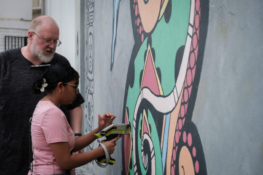 Two people using a device to test for lead in paint on a colorful mural featuring abstract art on a building wall.