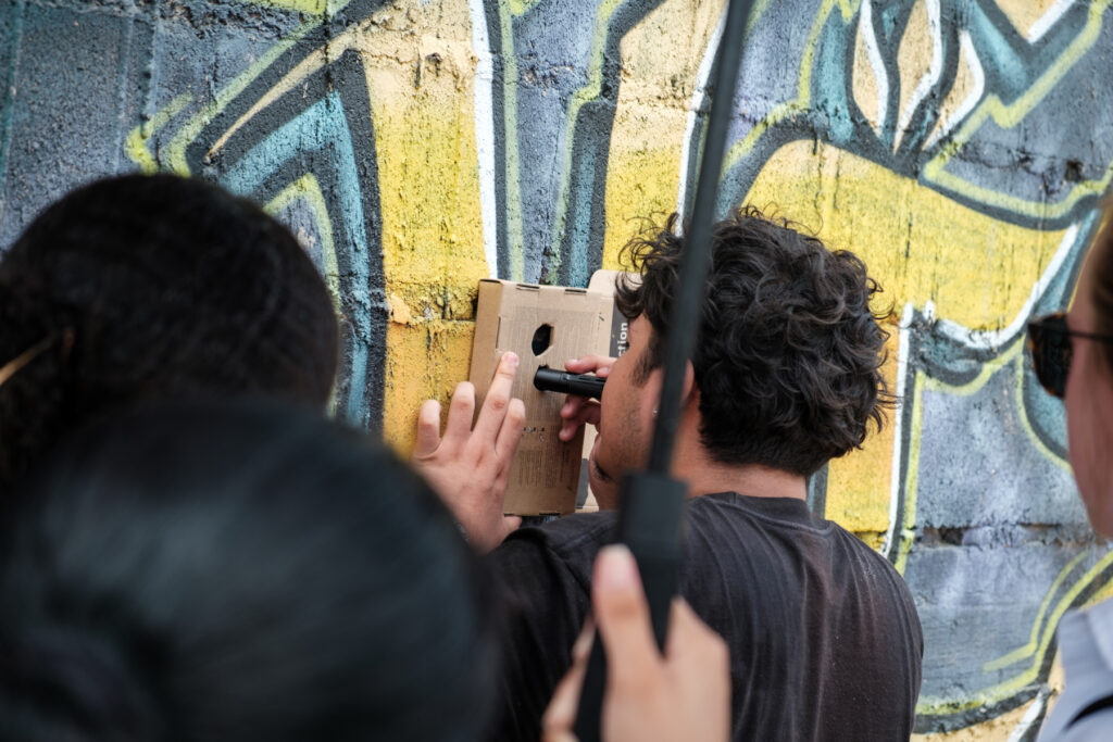 A group of people are gathered closely facing a graffiti-covered wall. One individual presses a small box with two holes against the wall and holds a small black cylinder inside the bottom hole.