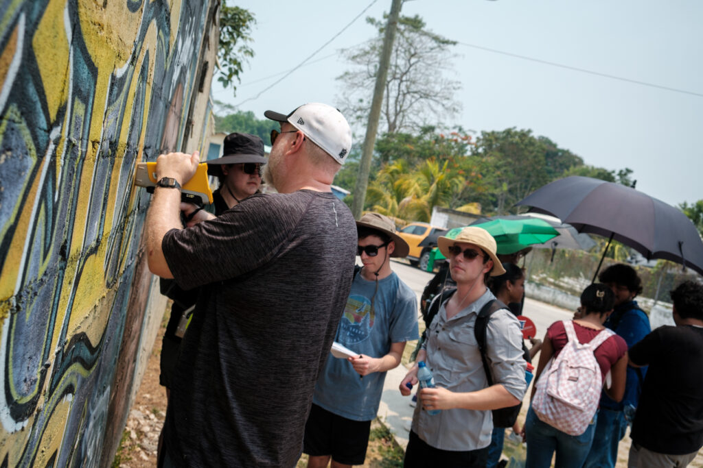 A group of college students gather around a colorful graffiti wall outside on a sunny day. Dr. Adam Kiefer is in the foreground pressing a device to the wall to test for lead in the paint while two students look on. Others in the background watch or protect themselves from the sun with umbrellas and hats.