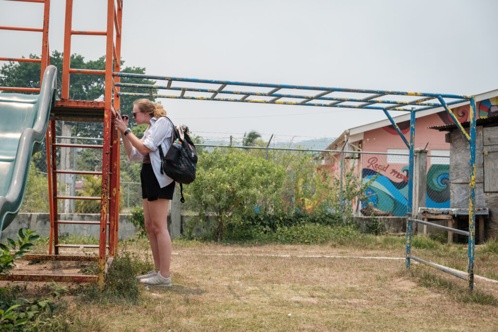 A Mercer University student with a backpack inspects a rusty, orange-painted ladder to the monkey bars at an outdoor playground. Colorful murals including wave patterns are visible on a surrounding building in the background.