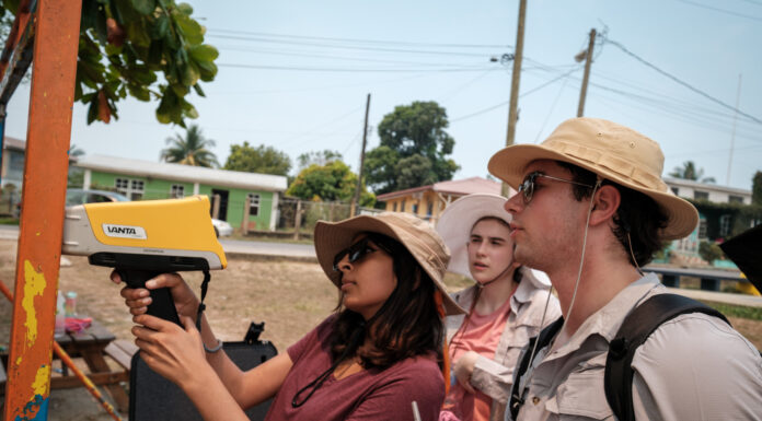 Mercer On Mission team tests for lead in paint, soil in Belize Three individuals use a pXRF device to test for lead on a metal bar with peeling orange paint outdoors. The person to the left operates the device as another takes notes, and a third observes. All are wearing sun hats.