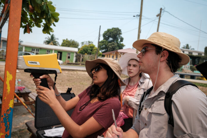 Three individuals use a pXRF device to test for lead on a metal bar with peeling orange paint outdoors. The person to the left operates the device as another takes notes, and a third observes. All are wearing sun hats.