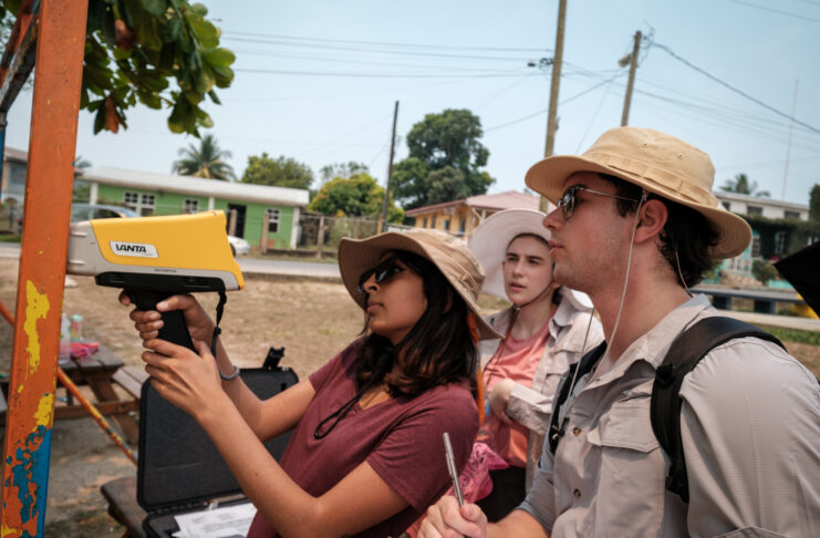 Three individuals use a pXRF device to test for lead on a metal bar with peeling orange paint outdoors. The person to the left operates the device as another takes notes, and a third observes. All are wearing sun hats.