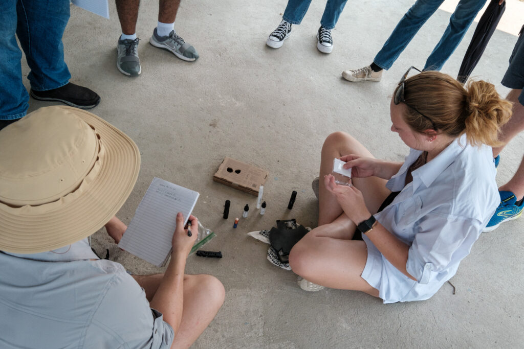 An individual sits on a concert floor with a small cardboard box and sprays in front of her. Others are gathered around watching and taking notes.