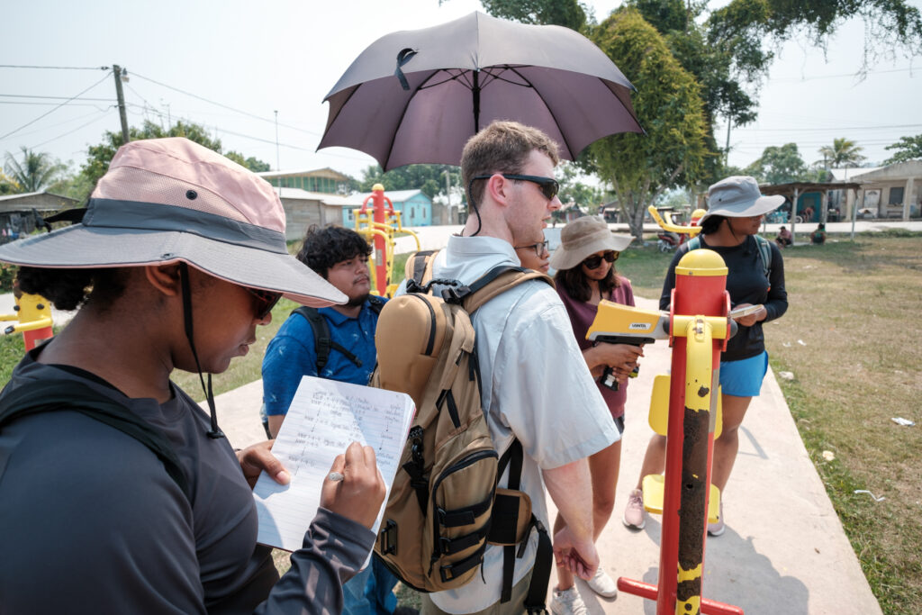 A group of college students are gathered around a rusted piece of outdoor exercise equipment. One individual is using a pXRF analyzer to test for lead and another is taking notes.