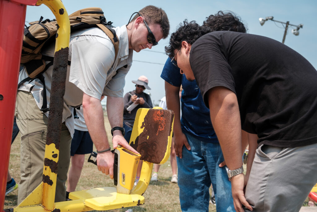 Two people use a handheld device to test the lead levels in paint on a piece of playground equipment, as other people do other work around them.