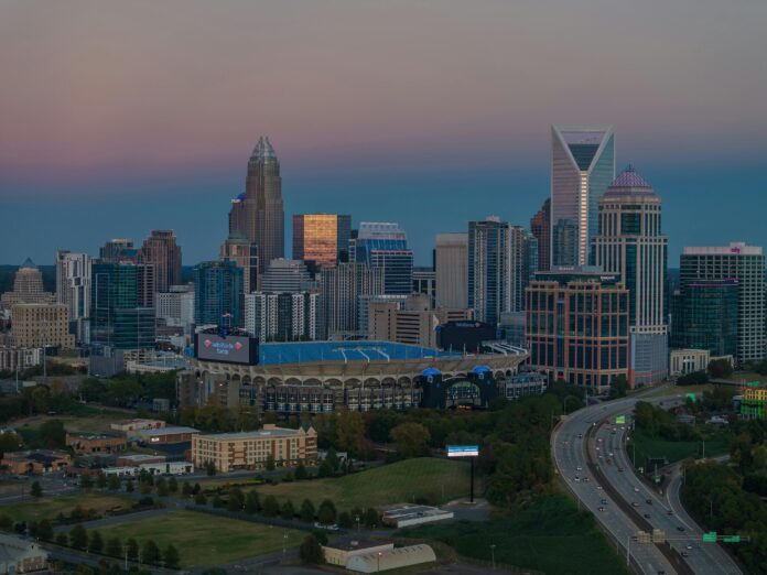 Dusk view of the Charlotte, North Carolina, skyline featuring modern skyscrapers with the Bank of America Stadium in the foreground and a highway leading into the city. The sky displays soft gradients from blue to pink.