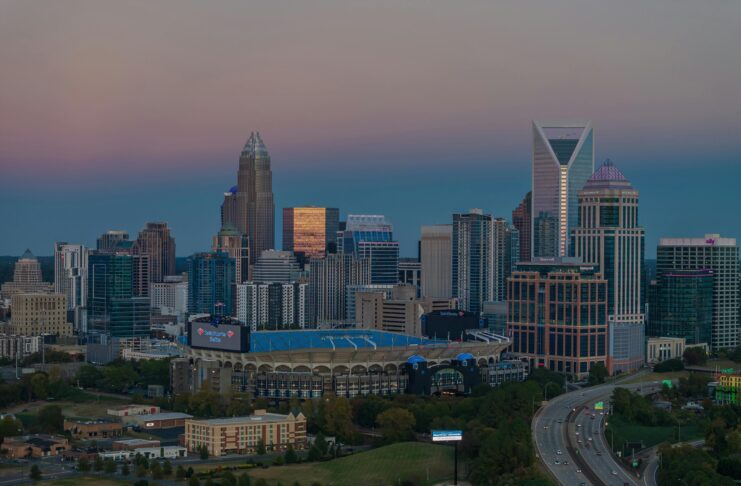 Dusk view of the Charlotte, North Carolina, skyline featuring modern skyscrapers with the Bank of America Stadium in the foreground and a highway leading into the city. The sky displays soft gradients from blue to pink.